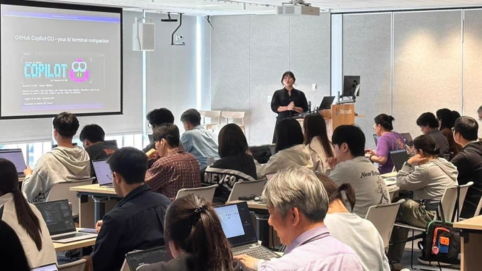Speaker presenting GitHub Copilot CLI to a seated audience in a bright classroom, with the Copilot CLI logo visible on the projection screen.
