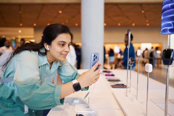 A person wearing AirPods and a green ombre blouse takes a photo of the AirPods display with iPhone 18 Pro in glacier.